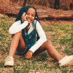 A girl sits in a forest in overalls, blue braids, and a big smile