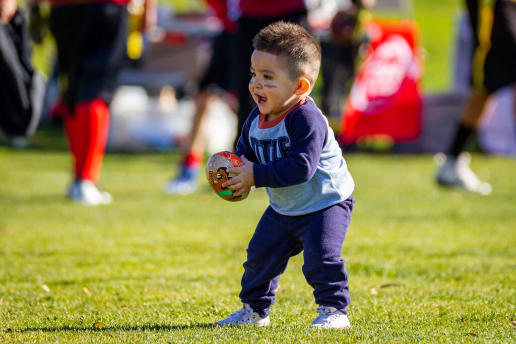 A toddler stands in a field holding a toy football and shouting joyfully