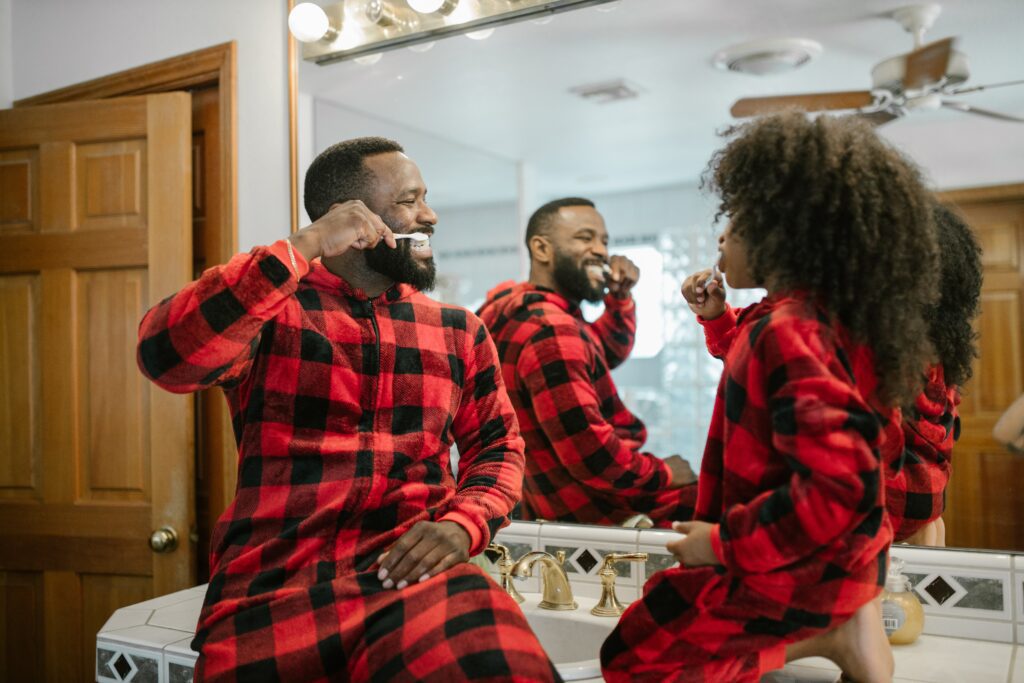 A father and child in matching buffalo plaid pajamas sit on the bathroom counter, smiling at each other as they both brush their teeth.