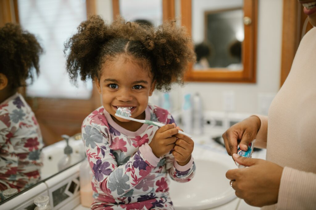 A toddler in a flowered shirt smiles and ponytails at the camera while brushing her teeth, as a parent stands nearby.