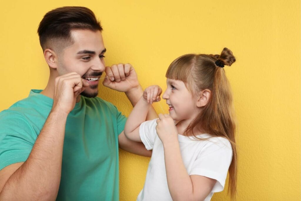 A young girl being taught how to floss her teeth by her dad